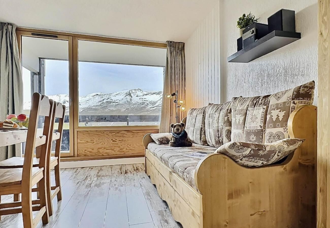En træsofa med mønstrede puder, et træbord med stole, og et stort vindue med udsigt til sneklædte bjerge.