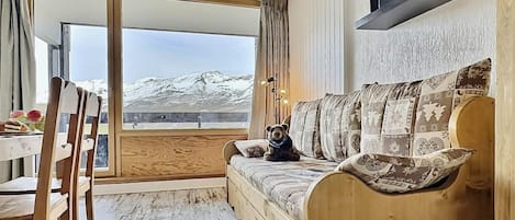 Un canapé en bois avec des coussins à motifs, une table en bois avec des chaises, et une grande fenêtre donnant sur des montagnes enneigées.