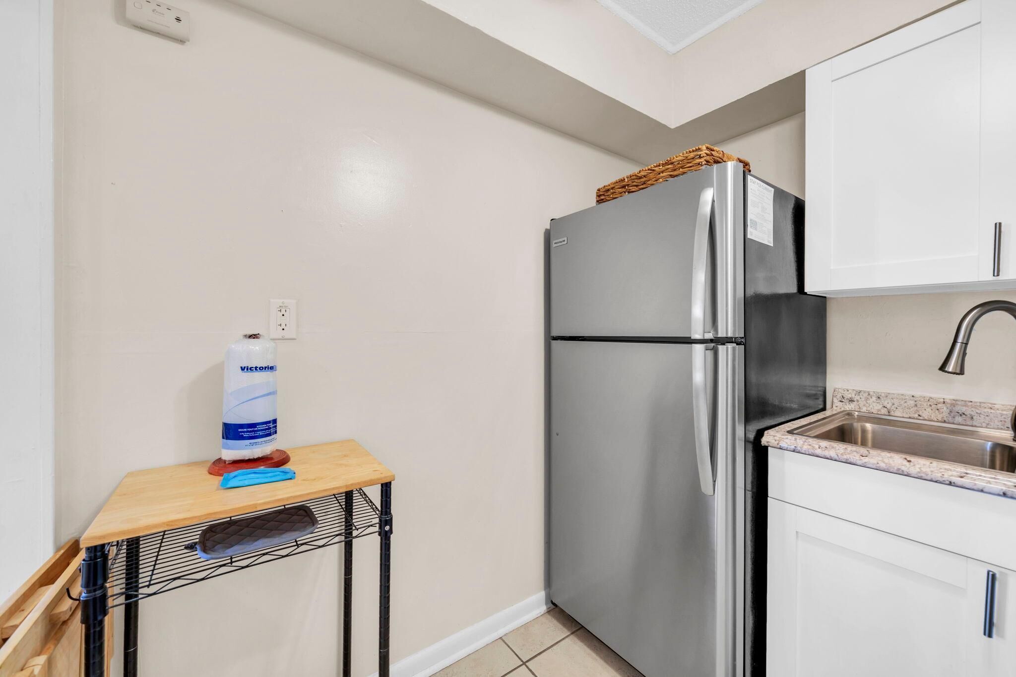 A compact kitchen with a stainless steel refrigerator, a sink, and a small wooden table with a bottle of hand sanitizer.