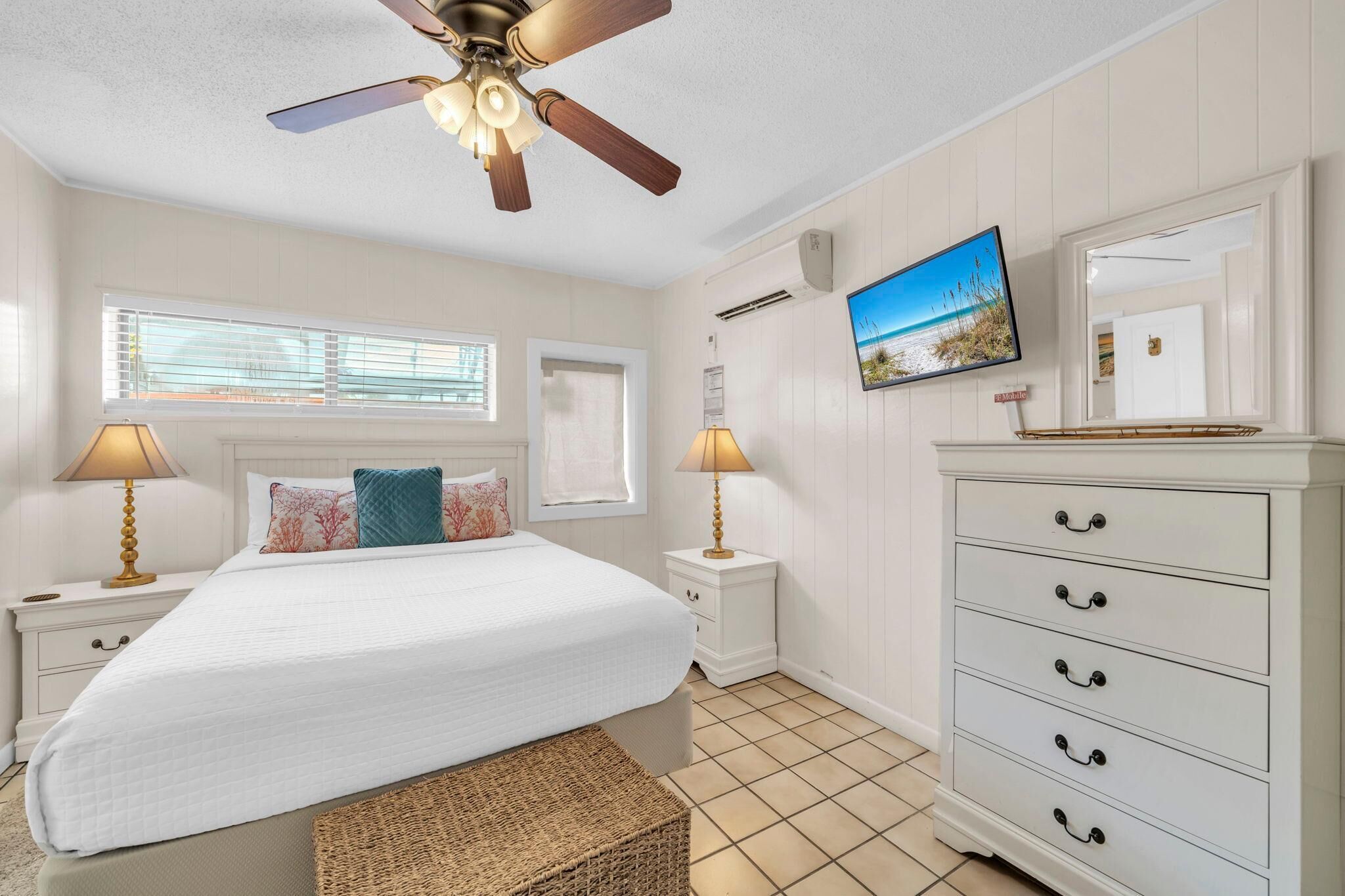 Ein Schlafzimmer mit einem Bett, Nachttischlampen, einem Schminktisch, einem Deckenventilator, einem Fernseher und einem Fenster mit Jalousien.