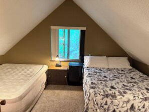 A small, triangular attic bedroom with a single window, a bedside table with a lamp, a mattress, and a bed with a patterned bedspread.