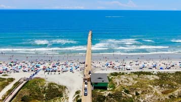 Une plage avec une longue jetée qui s’avance dans l’océan, bordée de monde et de parasols de plage.
