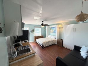 A compact room with a bed, a sofa, a kitchenette, and a ceiling fan.