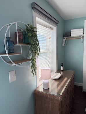 A room with a wicker table, a hanging plant, a window with blinds, and shelves with towels and jars.