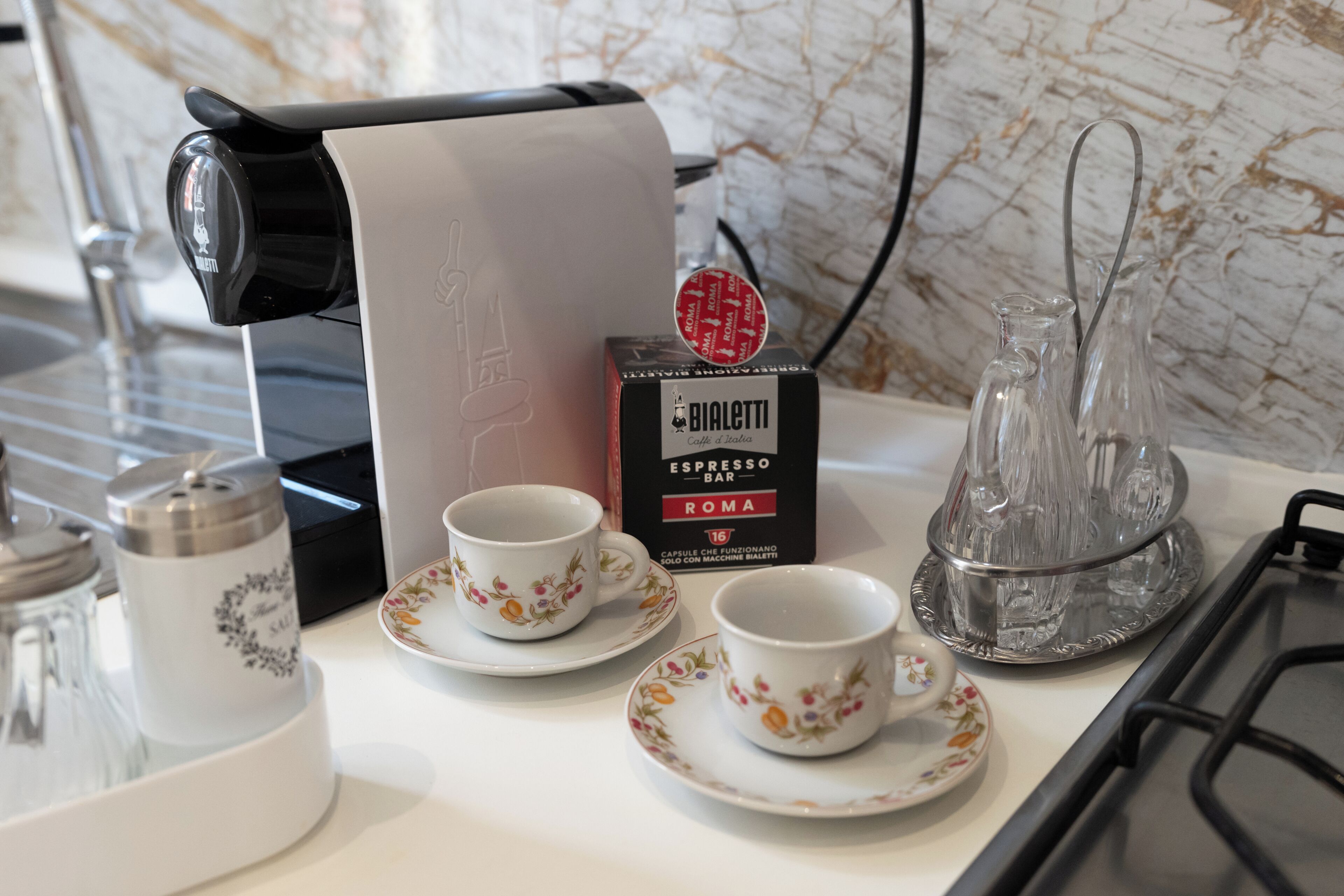 A kitchen counter with a coffee machine, two cups, a box of BIALETTI espresso bar, and salt and pepper shakers.