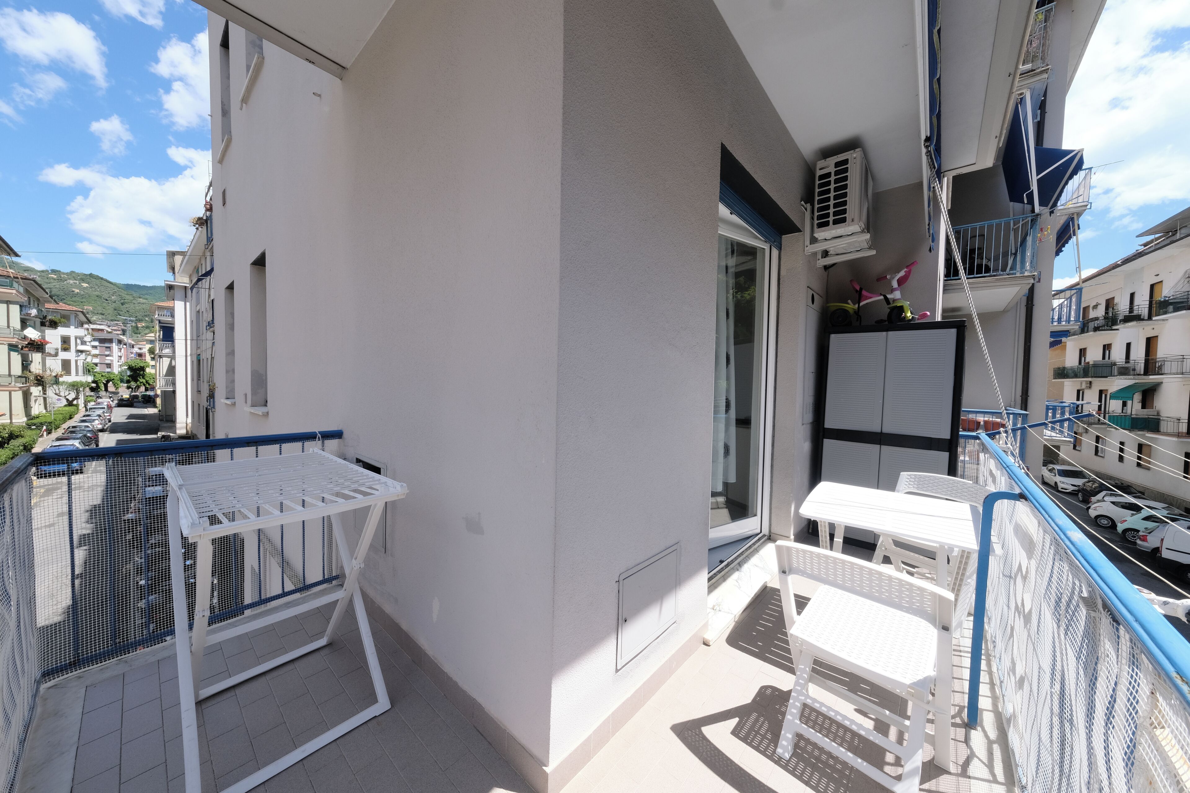 A balcony with a white table and chairs, a white metal grill, and a view of parked cars and buildings.