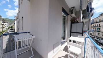 A balcony with a white table and chairs, a white metal grill, and a view of parked cars and buildings.