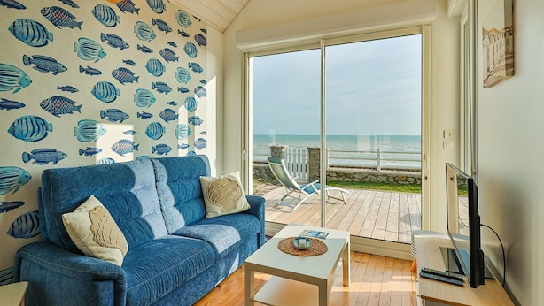 Una habitación con un sofá azul, una mesita blanca y un televisor. Se ve una ventana grande con vista al mar.