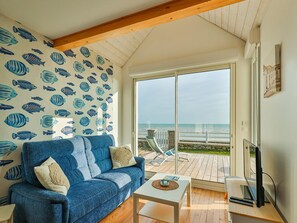 Quarto com sofá azul, uma pequena mesa branca e uma TV. Há uma janela grande com vista para o mar.