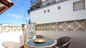 Een ronde glazen tafel met rieten stoelen op een terras, met een witte muur en decoratief lattenwerk.