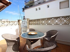 Ein runder Glastisch mit Korbstühlen auf einer Terrasse mit weißer Wand und dekorativem Gitter.
