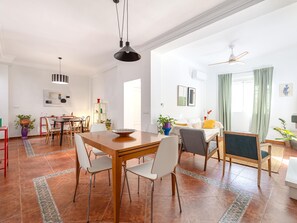 Ein geräumiger Wohnbereich mit Esstisch, Stühlen, einer Couch, Deckenventilator und einem großen Fenster mit Vorhängen.