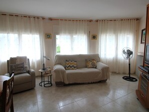 Ein Wohnzimmer mit Sofa, Sessel, kleinem Tisch und einem Standventilator.