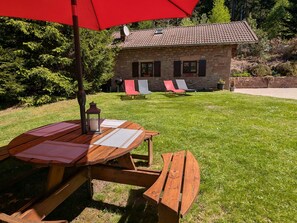 Un bâtiment en pierre avec un toit en tuiles, une table en bois surmontée d’un parasol rouge et deux chaises longues.