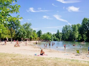 Una playa junto al lago con gente nadando y descansando, rodeada de árboles.