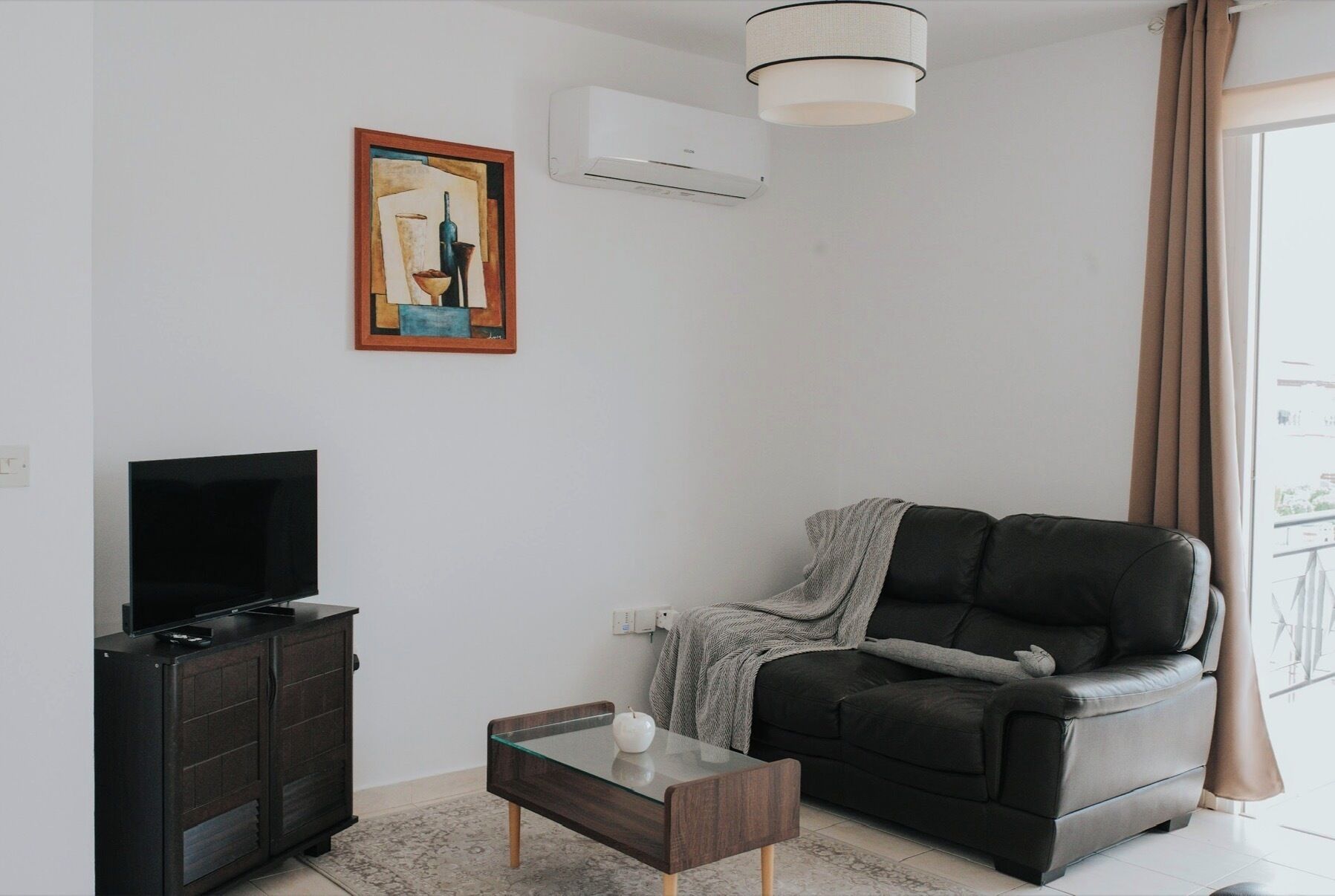 Ein Wohnzimmer mit einer Couch, einem Glastisch, einem Fernseher auf einem Schrank, einem Bild an der Wand und einer Klimaanlage.