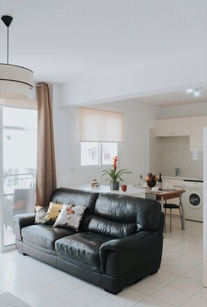 Ein modernes Wohnzimmer mit einer schwarzen Ledersofa, einem Esstisch mit Stühlen, einer Küchenzeile und einem großen Fenster mit Vorhängen.
