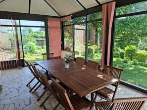 Een overdekt terras met een houten tafel en stoelen, grote ramen en uitzicht op een tuin.