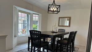 Un comedor con una mesa rectangular, seis sillas, una araña, una ventana con vista a un edificio de tejados rojos y un espejo en la pared.