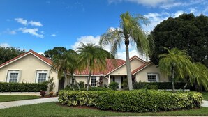 Una casa de dos pisos con techo de tejas rojas, rodeada de setos cuidados y palmeras.