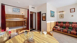Ein gemütliches Zimmer mit einem Etagenbett, einem Holzessttisch, einer gemusterten Couch und einer kleinen Küchenzeile.
