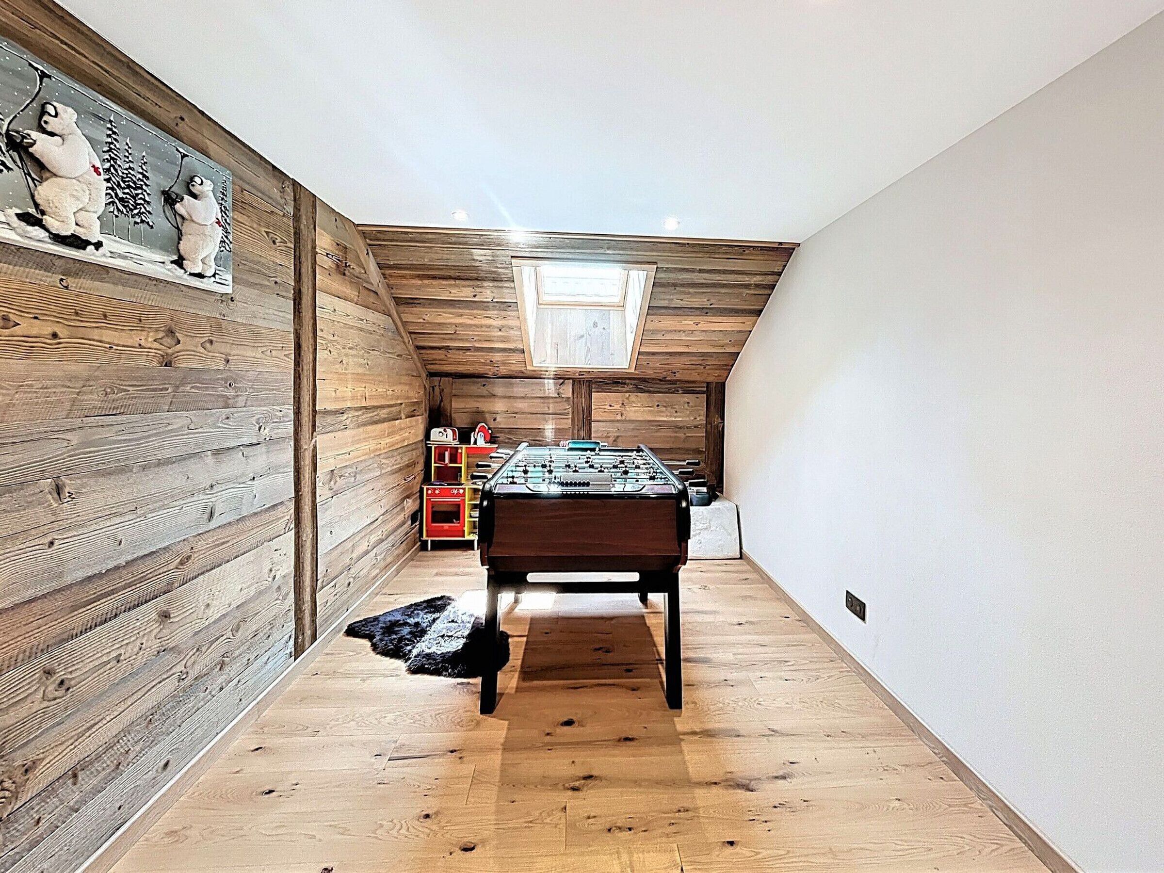 Um quarto de sótão estreito com paredes de madeira, uma mesa de pebolim e uma pequena janela.