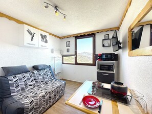 A cozy room with a sofa, a dining table with a red plate and cutlery, a microwave, and a window with a view of a mountain. - Studio mountain view - 2 guests (Le Corbier)