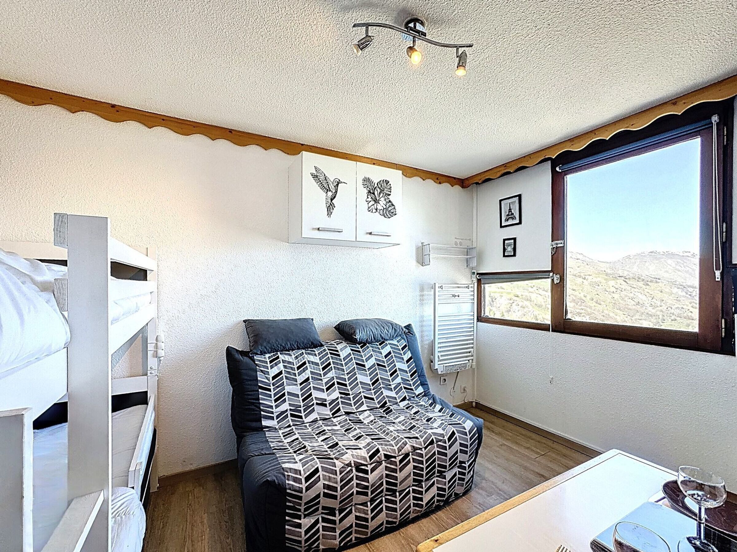 A room with a bunk bed, a sofa with patterned cushions, a window with a view of a hilly landscape, and wall-mounted artwork.