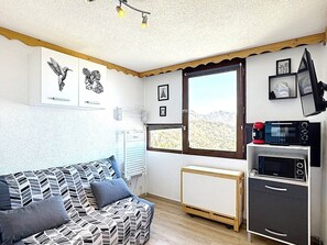 A compact room with a sofa, a small kitchenette with a microwave and coffee maker, a radiator, and a window with a view of a mountainous landscape. - Studio mountain view - 2 guests (Le Corbier)