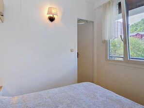 A room with a bed, a wall-mounted light fixture, a door, and a window with a view of greenery and buildings. - Bright apartment in the heart of the slopes - 6 guests (La Toussuire)
