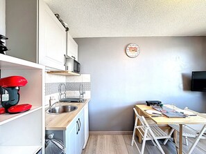 A compact kitchen with a sink, microwave, and red appliance on the left. A dining table with chairs is on the right, with a clock on the wall. - Studio with mountain view, ski-in/ski-out - 2 guests (Le Corbier)