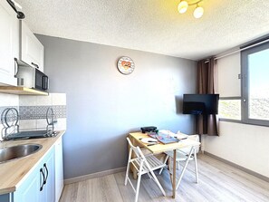 A compact kitchen with a microwave, sink, and a clock on the wall. There's a dining area with a table and chairs, and a TV mounted on the wall. - Studio with mountain view, ski-in/ski-out - 2 guests (Le Corbier)