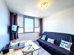 A room with a dark blue sofa, a flat-screen TV mounted on the wall, a dining table with a placemat, and a window with curtains. - Studio with mountain view, ski-in/ski-out - 2 guests (Le Corbier)