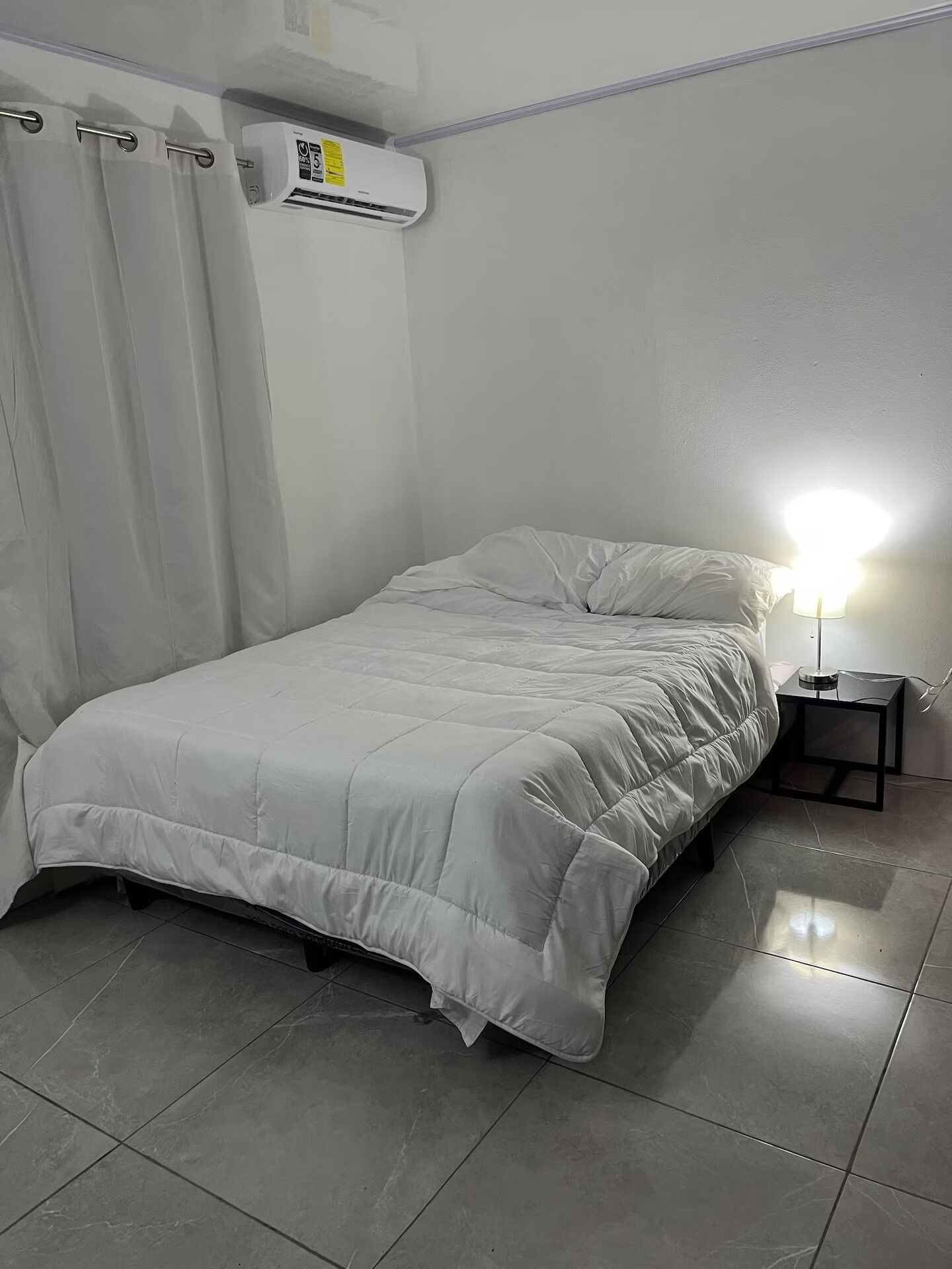 Ein Schlafzimmer mit einem Bett, einem Nachttisch, einer Lampe, einem Fenster mit Vorhängen und einer Klimaanlage.
