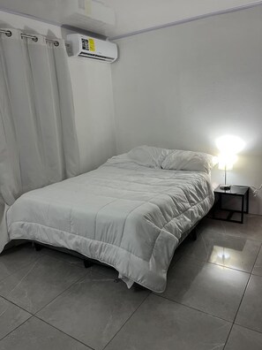 Ein Schlafzimmer mit einem Bett, einem Nachttisch, einer Lampe, einem Fenster mit Vorhängen und einer Klimaanlage.