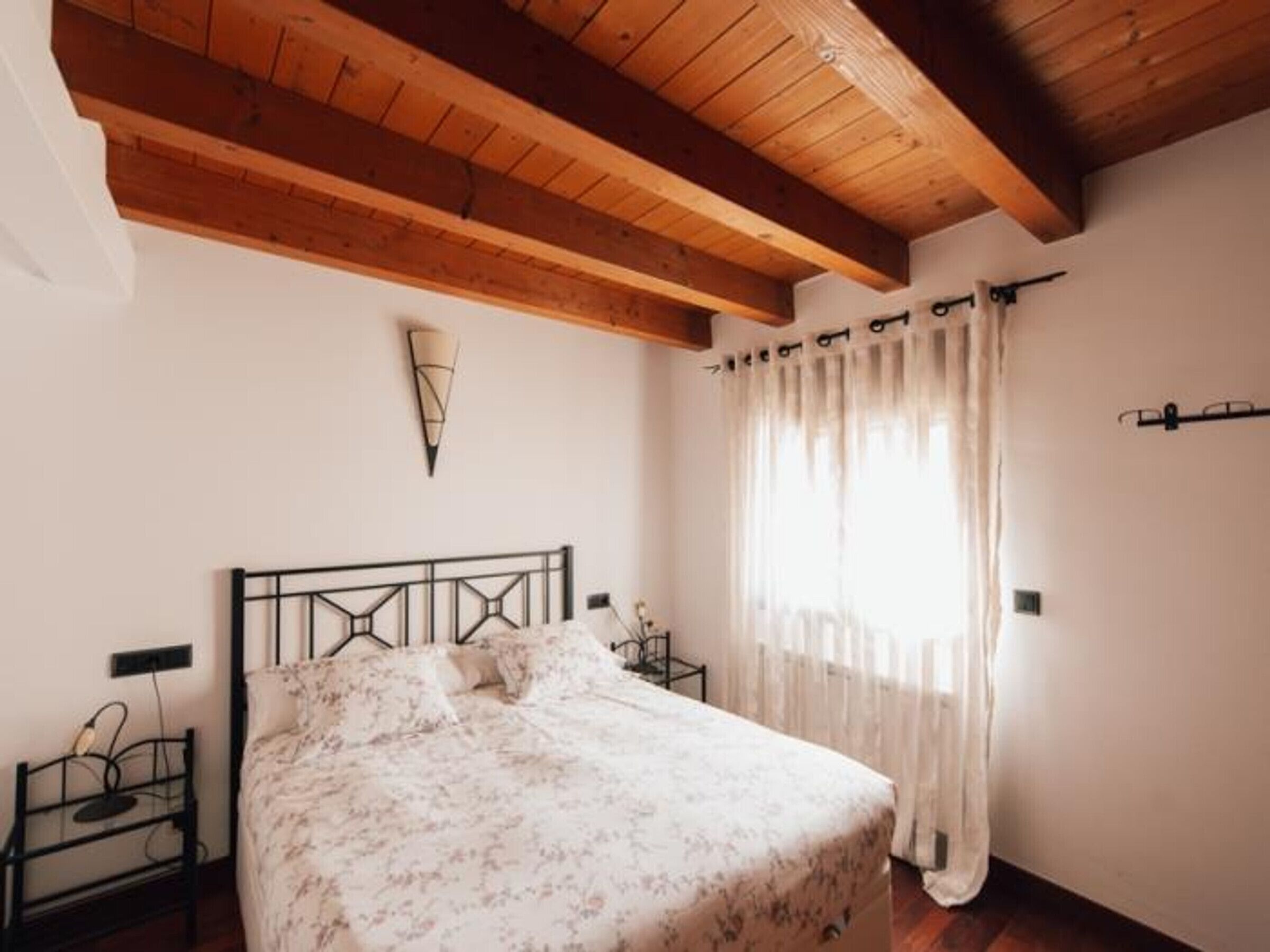 Ein Schlafzimmer mit Holzboden, einem Bett mit floralem Überwurf, einem Nachttisch mit Lampe und einem Fenster mit transparenten Vorhängen.