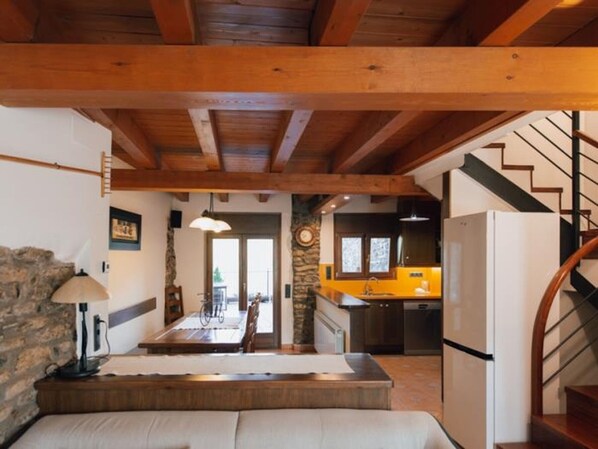 A rustic interior with exposed wooden beams, a stone wall, a dining area, and a kitchen with a yellow countertop.