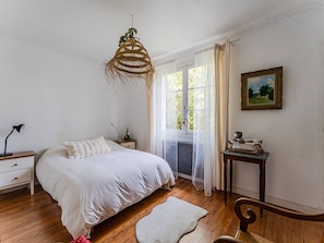 Ein Schlafzimmer mit einem Bett, einem Schreibtisch, einem Stuhl, einer Pendelleuchte, einem Fenster mit Vorhängen und einem gerahmten Bild an der Wand.