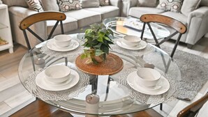 Une table ronde en verre, surmontée de quatre assiettes et bols blancs, est entourée de chaises à ossature en bois. L’ensemble est disposé dans un salon où se trouvent un canapé gris et des coussins à motifs.