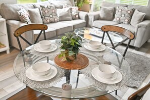 A round glass table with four white plates and bowls, surrounded by chairs with wooden frames, set in a living room with a grey sofa and patterned cushions. - Disney Dream~Resort Pool & Water Park Fun~Hot Tub (Kissimmee)