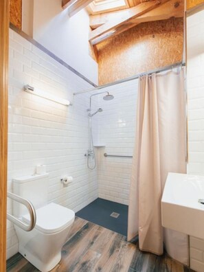 Une salle de bain avec des murs carrelés de carreaux blancs, un plancher en bois, une douche avec un rideau, des toilettes équipées d’une barre d’appui et un lavabo avec un miroir.
