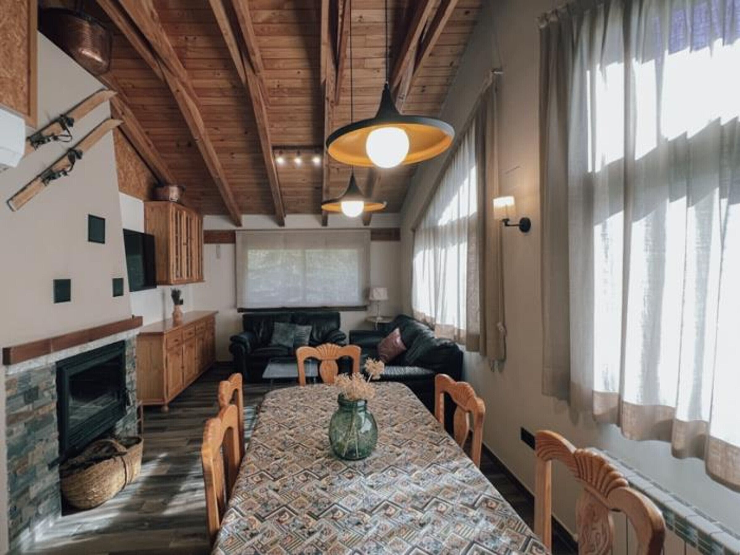 Ein gemütliches Wohnzimmer mit Kamin, Holzbalkendecke, einem Esstisch mit Stühlen und einem großen Fenster mit Vorhängen.