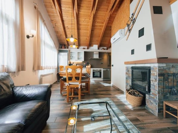 A cozy living room with a leather sofa, a glass coffee table, wooden dining table, and chairs. A fireplace is visible in the background.