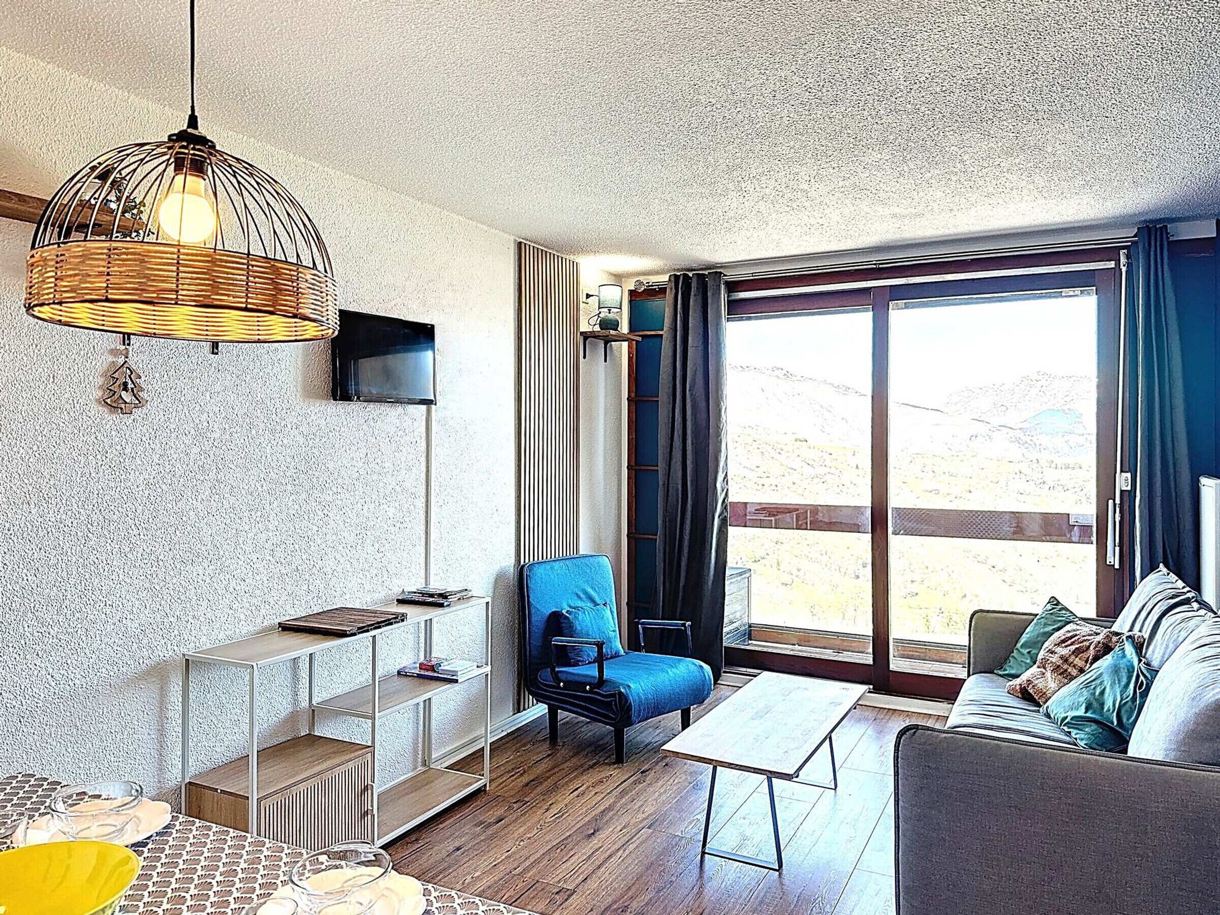 A modern living room with a sofa, a coffee table, a TV mounted on the wall, a wicker light fixture, and a large window with a view.