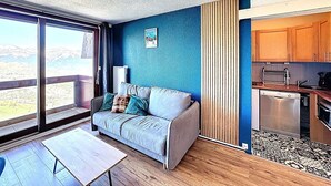 A modern room with a sofa, a table, a view through a large window, and a kitchenette. - Studio ski-in/ski-out, valley view - 5 guests (Le Corbier)
