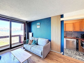 A modern room with a sofa, a table, a view through a large window, and a kitchenette. - Studio ski-in/ski-out, valley view - 5 guests (Le Corbier)