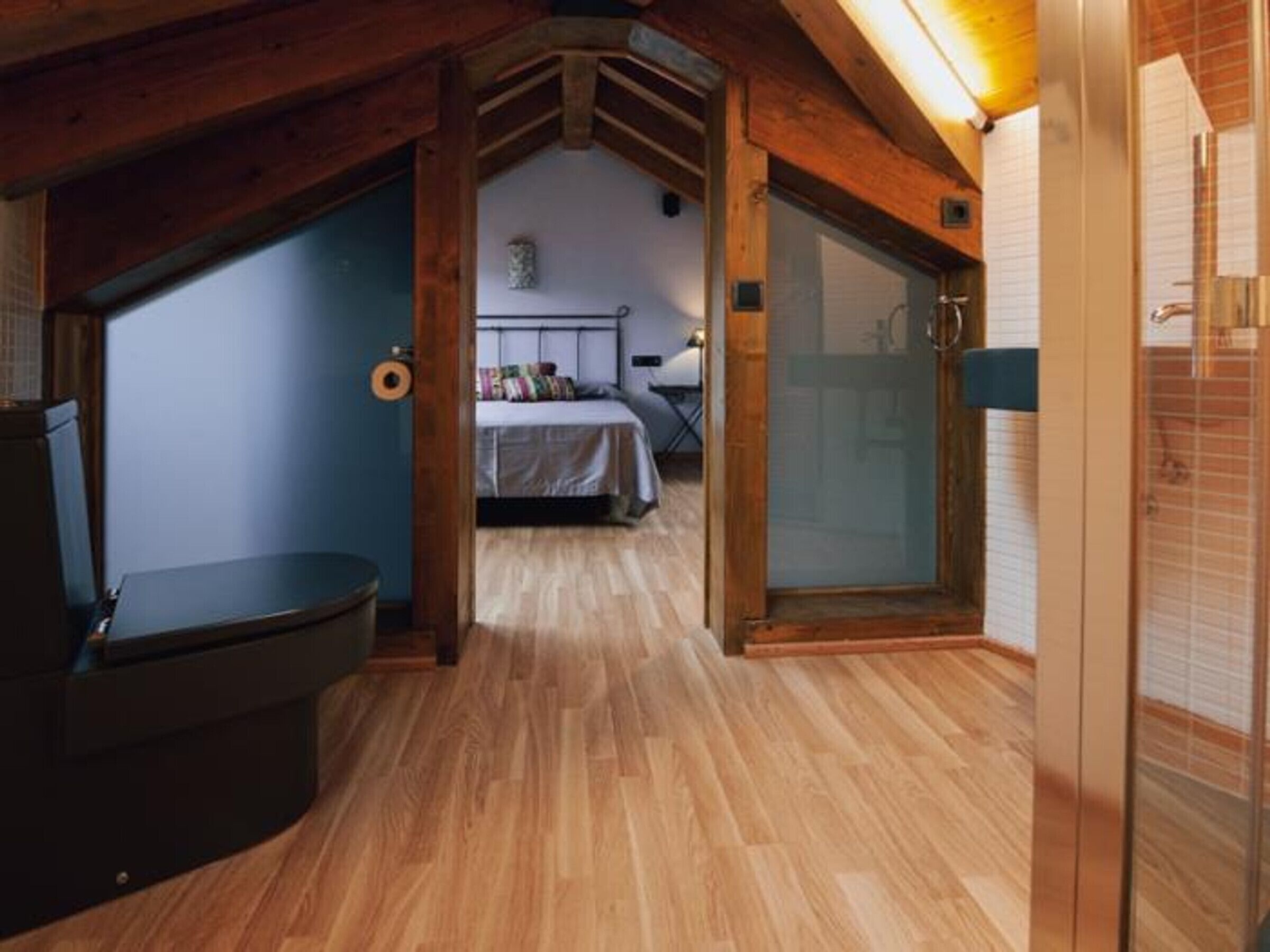 Un pasillo estrecho con suelo de madera, una cama con cabecera colorida y una puerta de cristal que da a un baño.