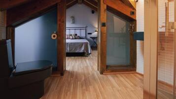 Un pasillo estrecho con suelo de madera, una cama con cabecera colorida y una puerta de cristal que da a un baño.