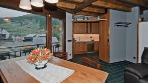 A wooden dining table with a lace table runner and a vase of flowers, a leather sofa, and a kitchen area with wooden cabinets and a view of a village through large windows. - Cozy House near Aigüestortes Park, 6 ppl, Pets welcome (Espot)