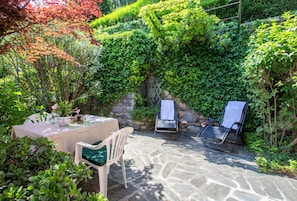A patio with a table, chairs, and a stone floor surrounded by lush greenery and a stone wall.